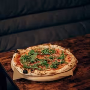 A freshly baked wood-fired pizza topped with arugula served on a rustic wooden table.